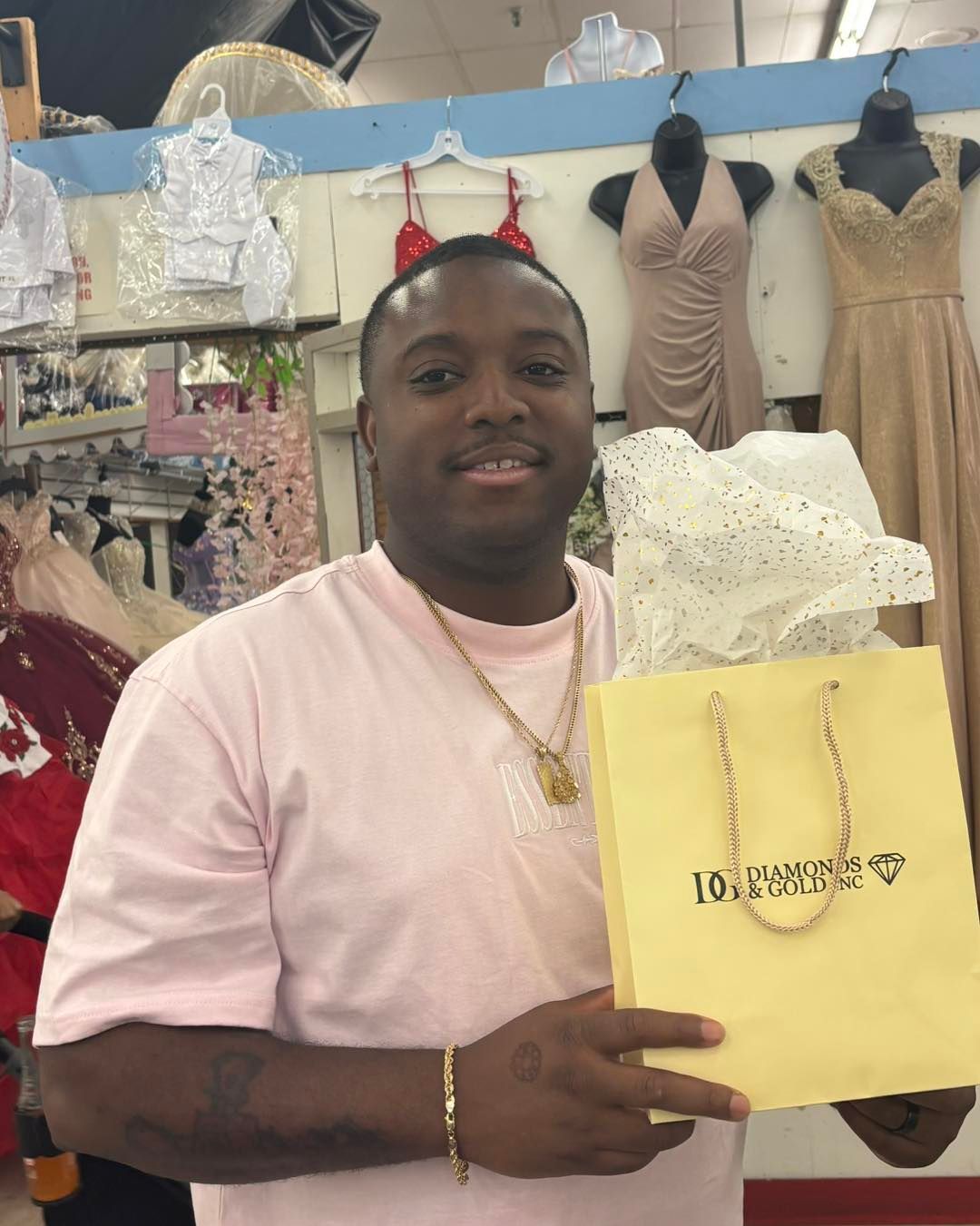 Man smiling, holding a gold gift bag at a dress shop, dresses visible in background.