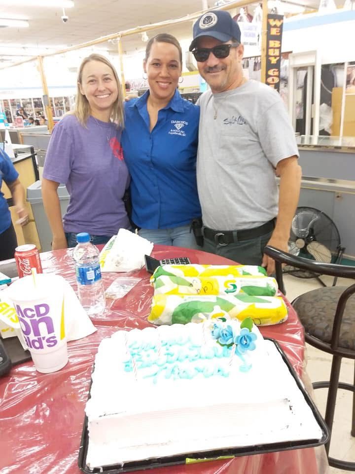 Three people smile around a cake. A red tablecloth is on a table with food.