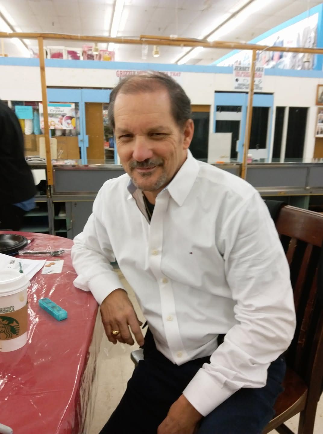 Man in white shirt smiling, seated at a table. Indoor setting with shelving visible in the background.