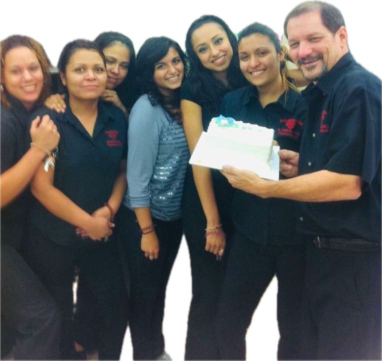 Group of people smiling, holding a cake. Dark clothing, light skin tones. Indoor setting.