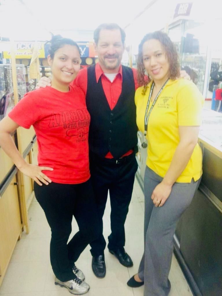 Three people pose in a store aisle. Woman in red shirt and black pants, man in vest, and woman in yellow shirt and gray pants.
