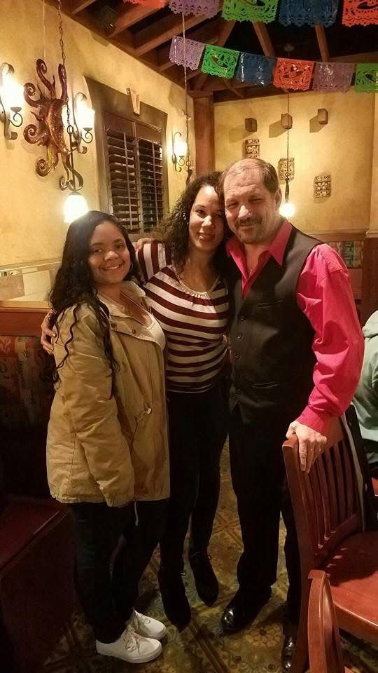 Three people smiling, posing together in a decorated restaurant. One wears a pink shirt, vest. The other two, a jacket, and a striped shirt.