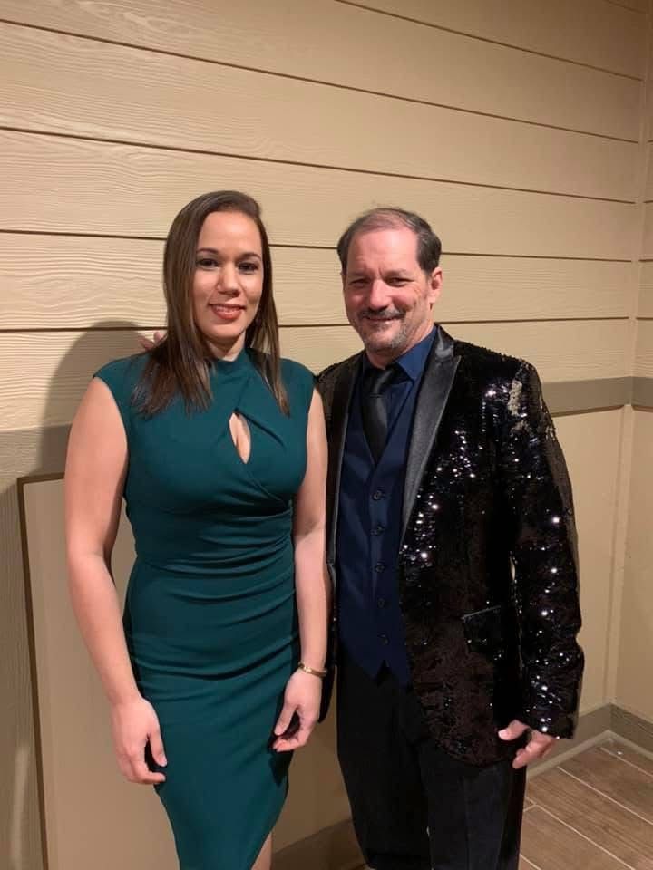 Woman in teal dress and man in sequin jacket pose together in front of a wooden wall.