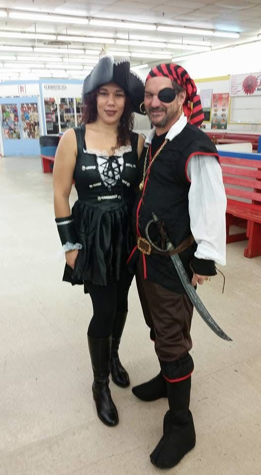 Two people in pirate costumes, posing indoors. Woman wears a black dress, hat, and boots. Man has an eye patch and sword.