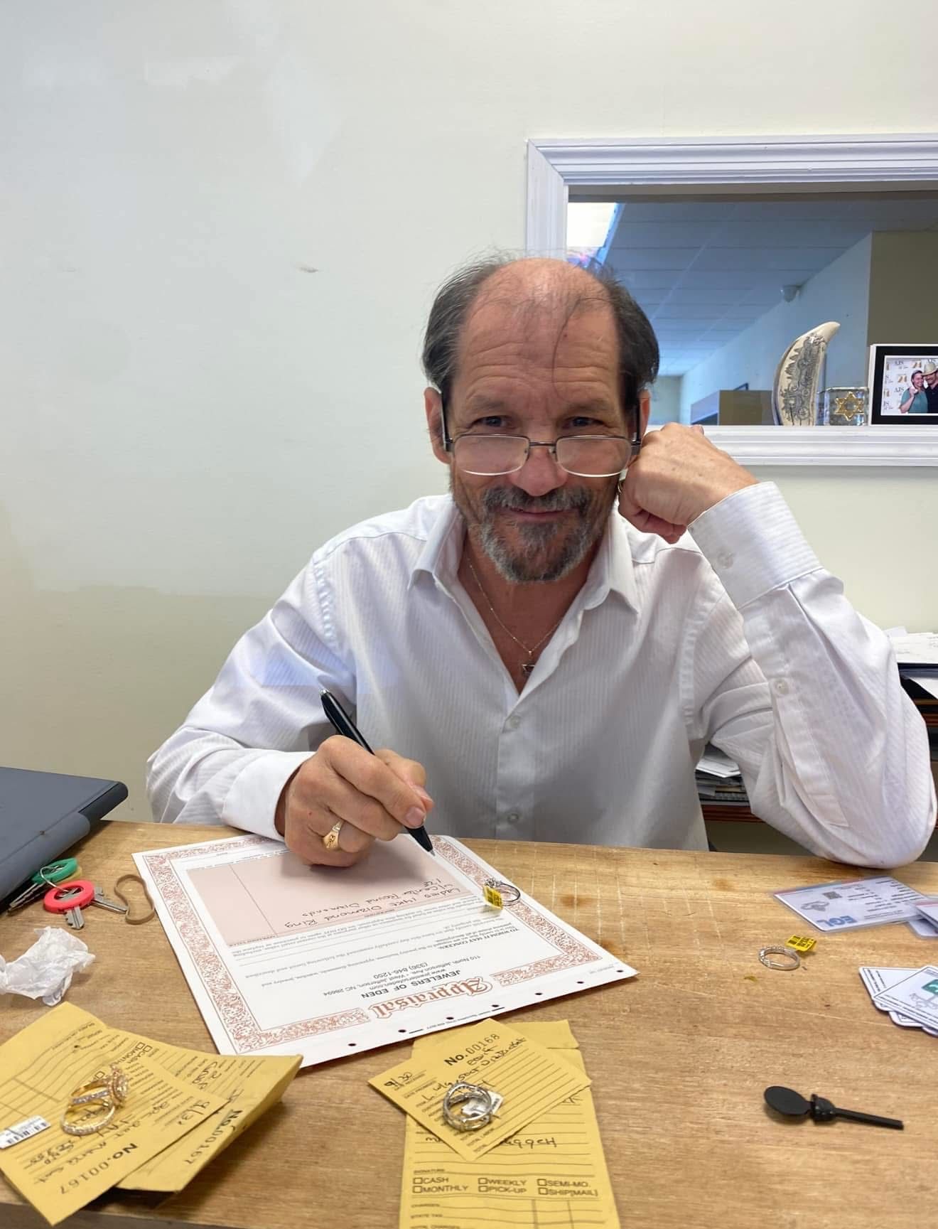 Man signing document at desk, looking at the camera. Papers, desk tools and accessories are present.