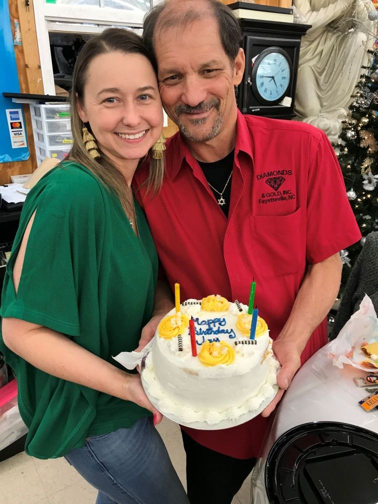 Woman and man holding birthday cake, both smiling. Cake has candles and