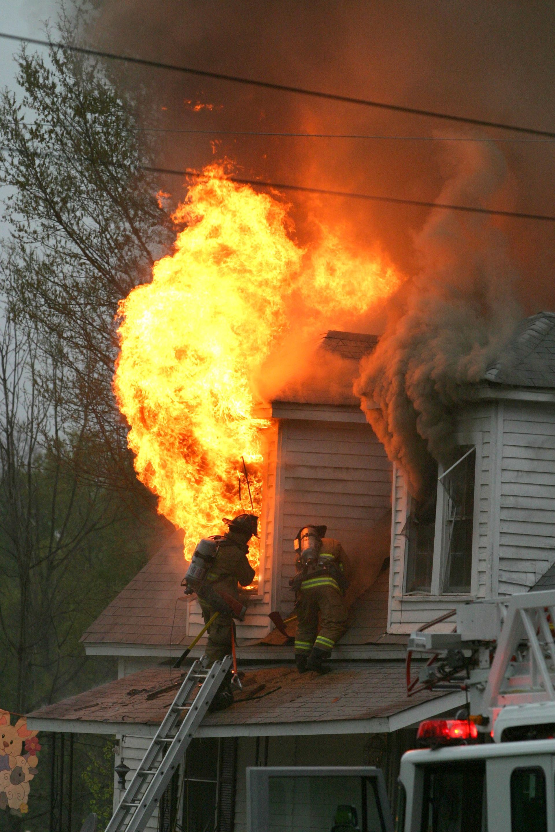 Firefighters on ladder battling house fire; flames and smoke billowing.