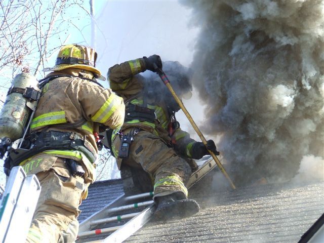 Firefighters on roof with smoke, one uses a tool, another stands by, ladder visible.