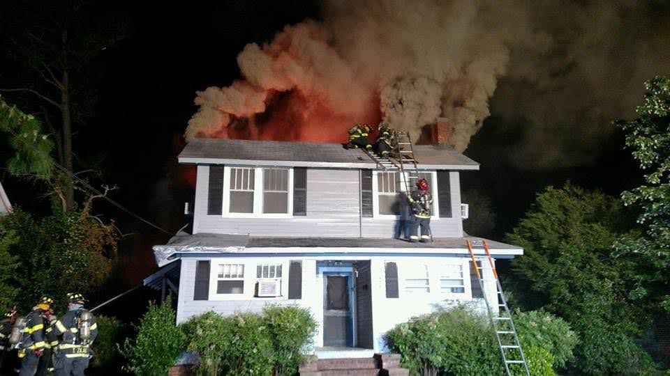 House engulfed in flames at night with firefighters on the roof and a ladder.