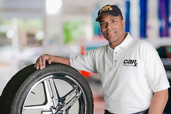 Man in a cap and shirt leans on a tire in a car shop, smiling.