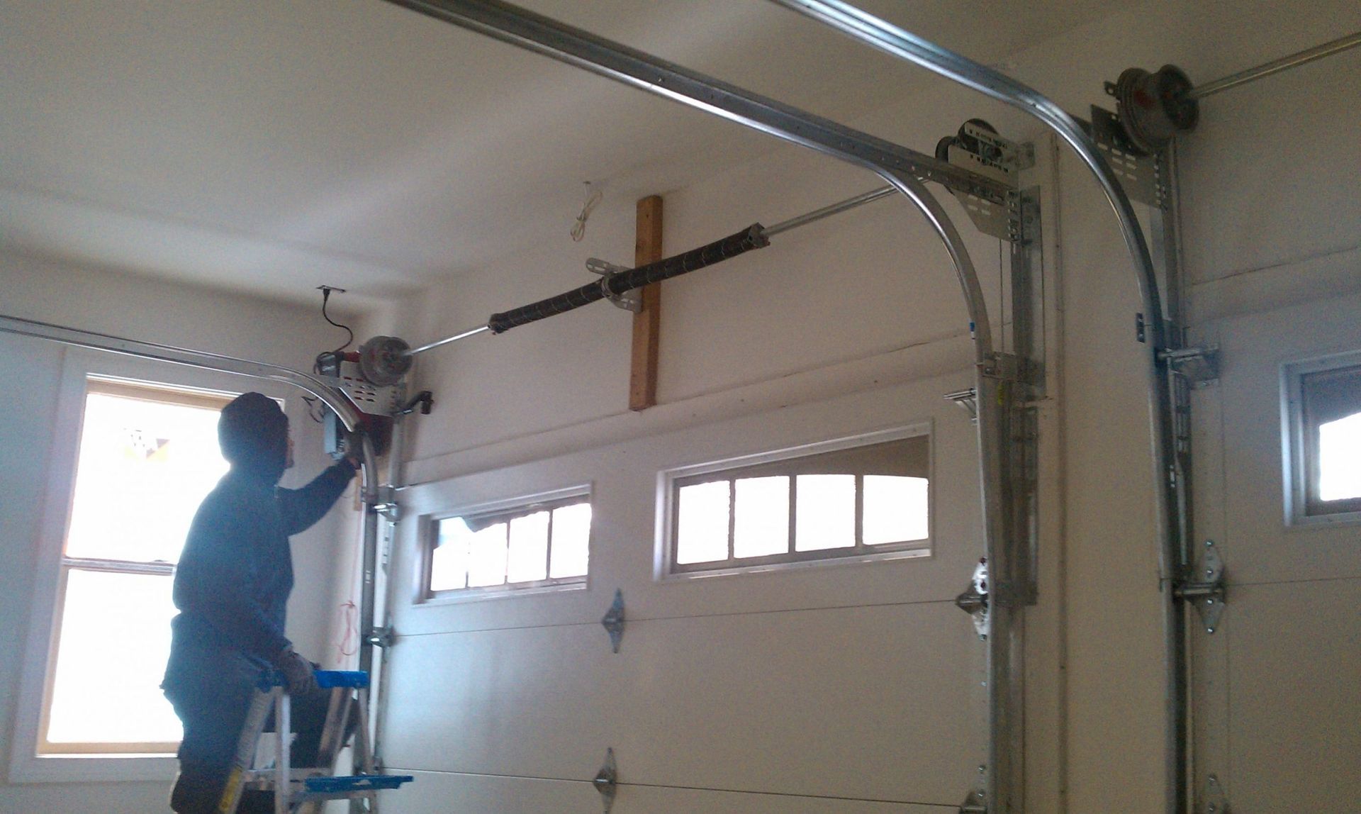 Man on a ladder repairing a garage door spring. Indoors, white garage door.