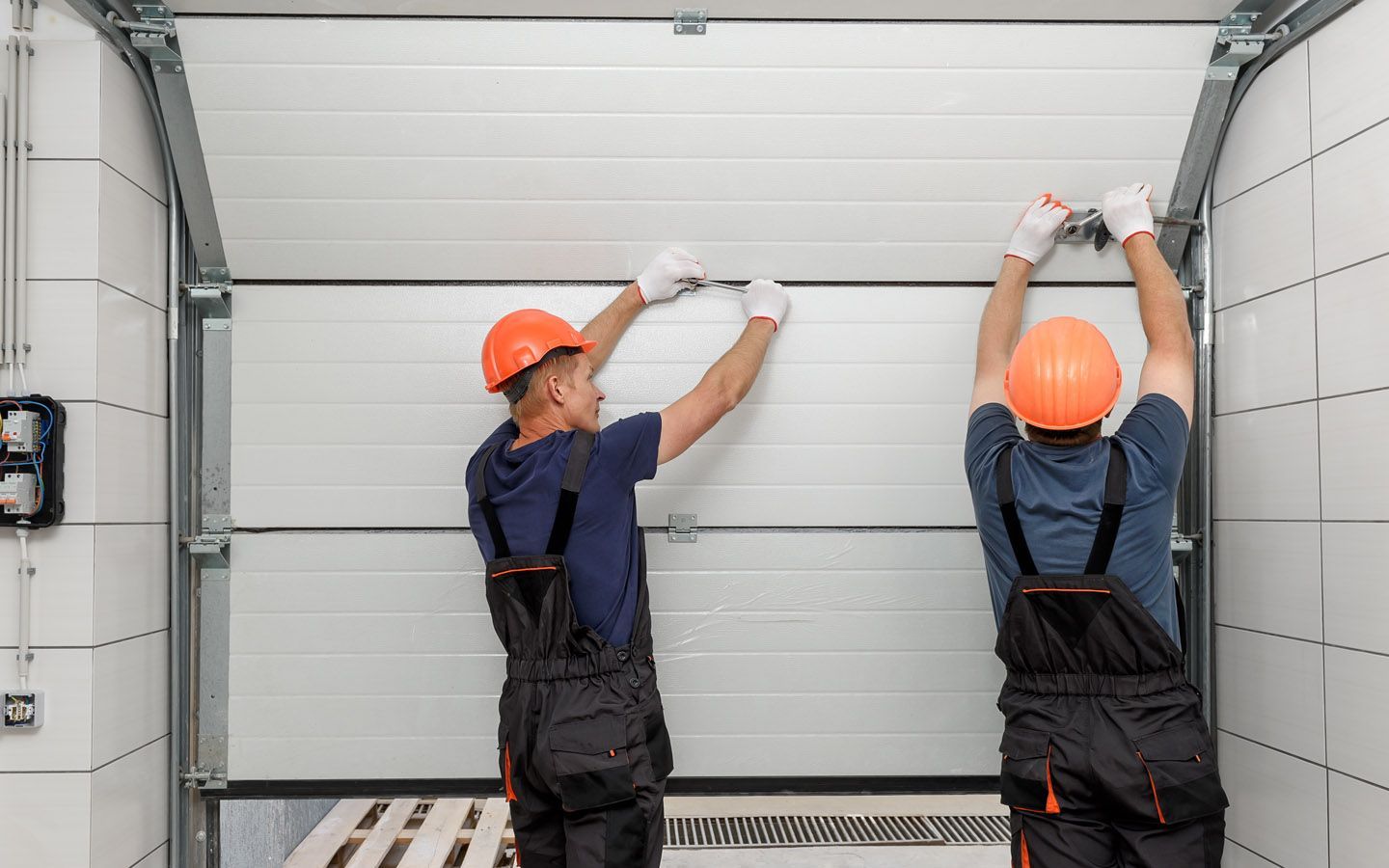Two workers in overalls and hard hats install a white garage door.