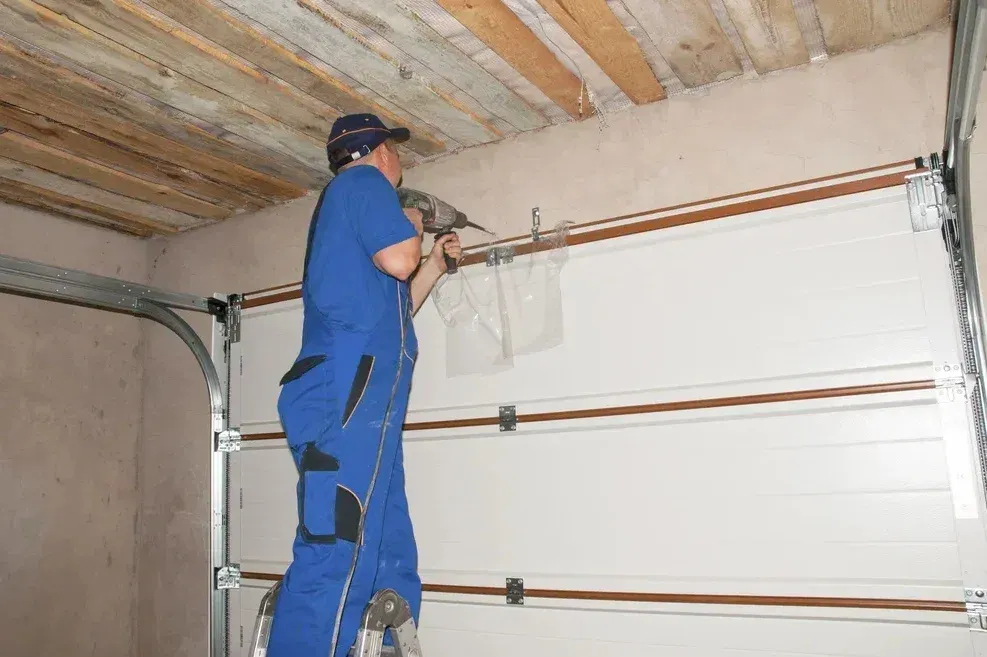 Man in blue overalls on ladder, installing garage door. Interior shot.