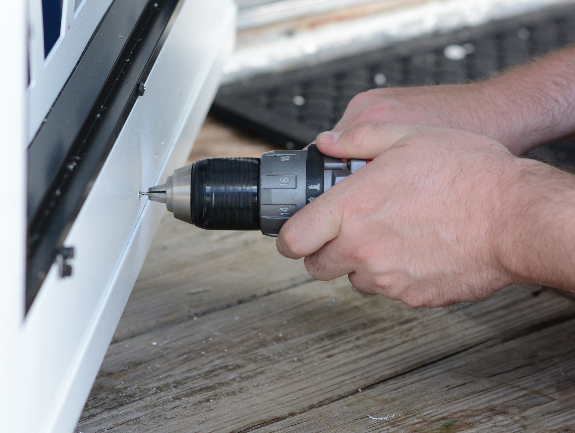 Person using a drill to secure a white panel; outdoors on a wooden surface.