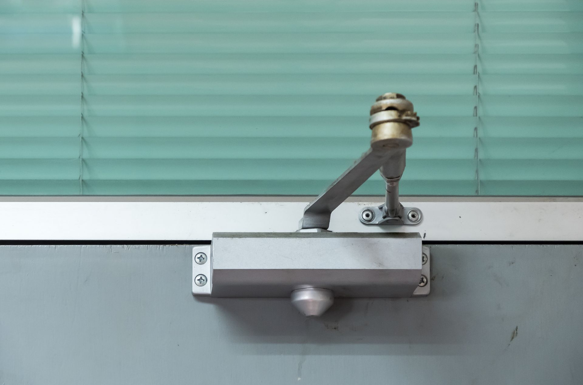 Silver door closer on a gray door, connected to an arm and mounted near a window with blinds.
