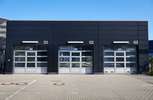 Three garage doors with windows, numbered 01-03, under a black building exterior, on a sunny day.