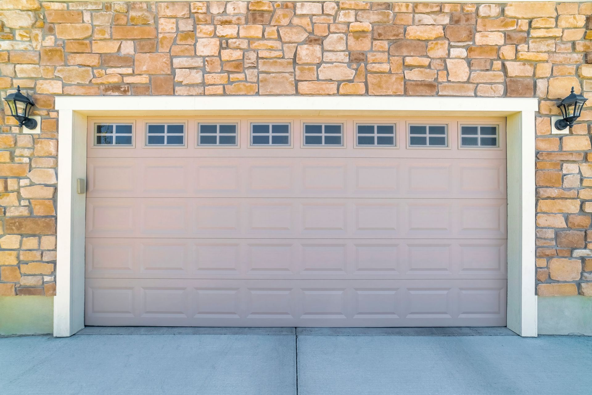 Beige garage door with small square windows, set in a stone wall, two black sconces on either side.