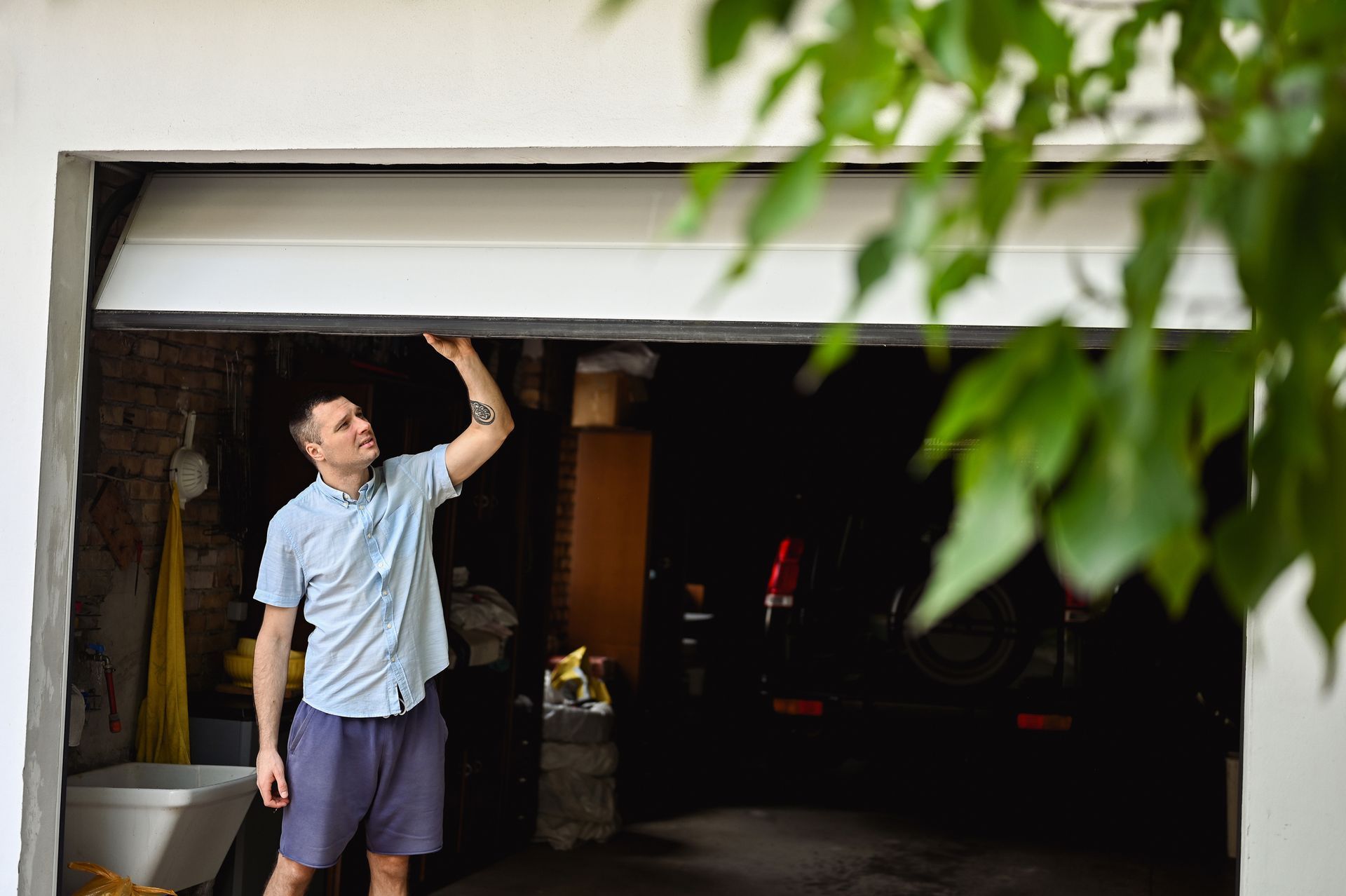 Man in light blue shirt reaching up to open a garage door, outdoors.