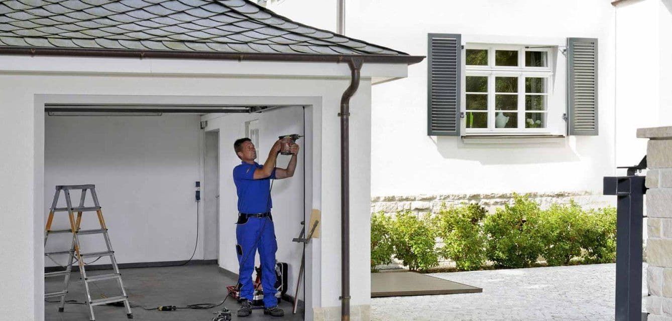 Man in blue jumpsuit drilling in a garage, with a ladder, white building, and shutters.