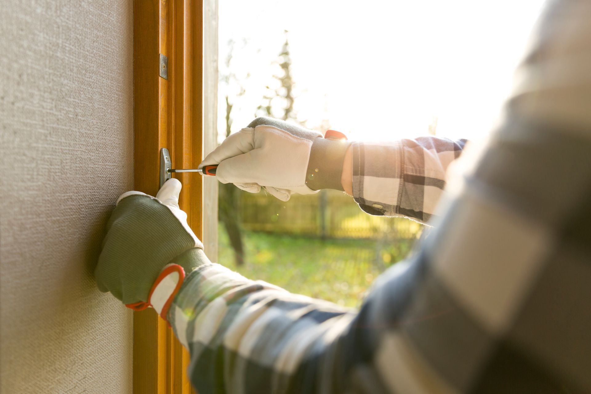 Person in plaid shirt using a screwdriver on a screen door, wearing gloves. Sunlight illuminates the outdoor setting.