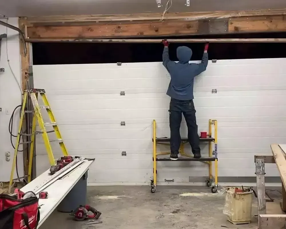 Person installing a white garage door, standing on a yellow scaffold in a garage.