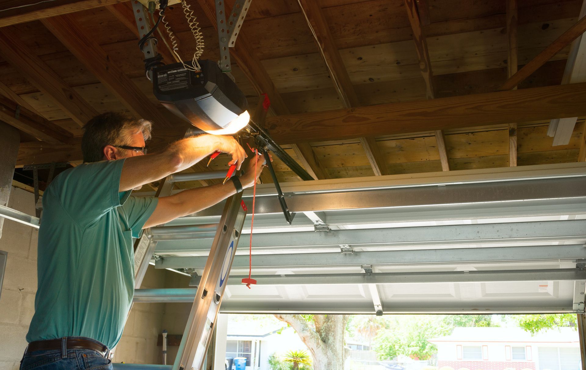 Man on ladder working on garage door opener. Inside a garage with sunlight.