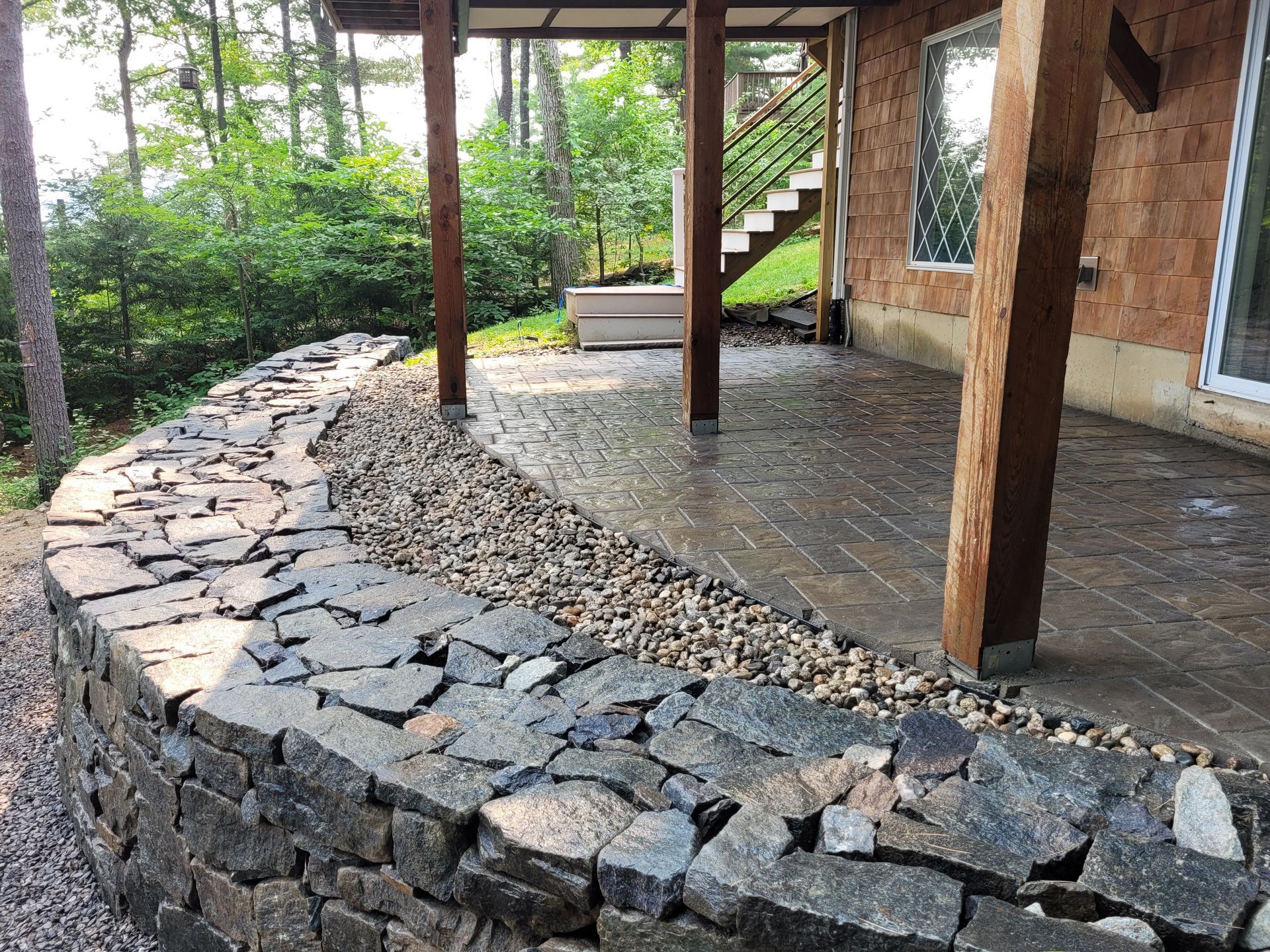 A curved, dark stone retaining wall borders a stone patio and wooden porch posts against a house, with trees in the back.