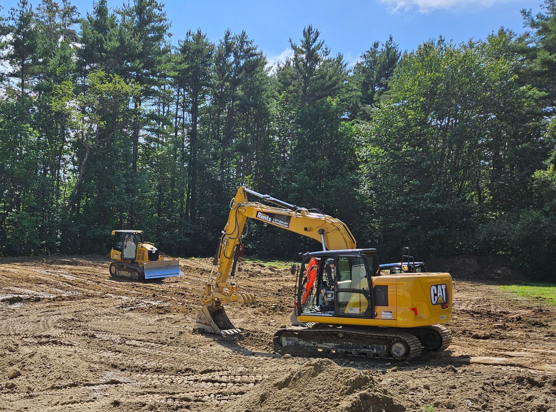 A yellow excavator rests on a dirt construction site against a backdrop of trees under a clear blue sky.