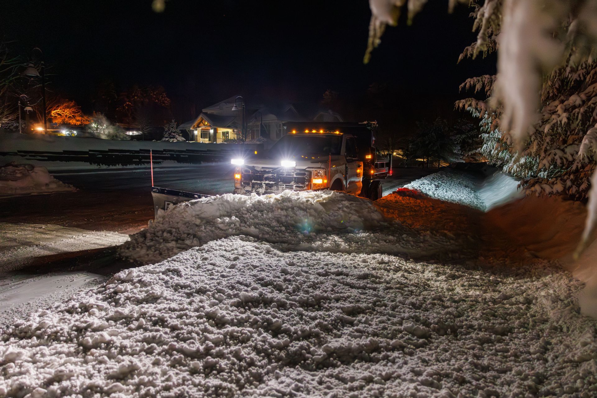 A snowplow clears a snow-covered residential driveway at night, its bright headlights illuminating the falling snow.