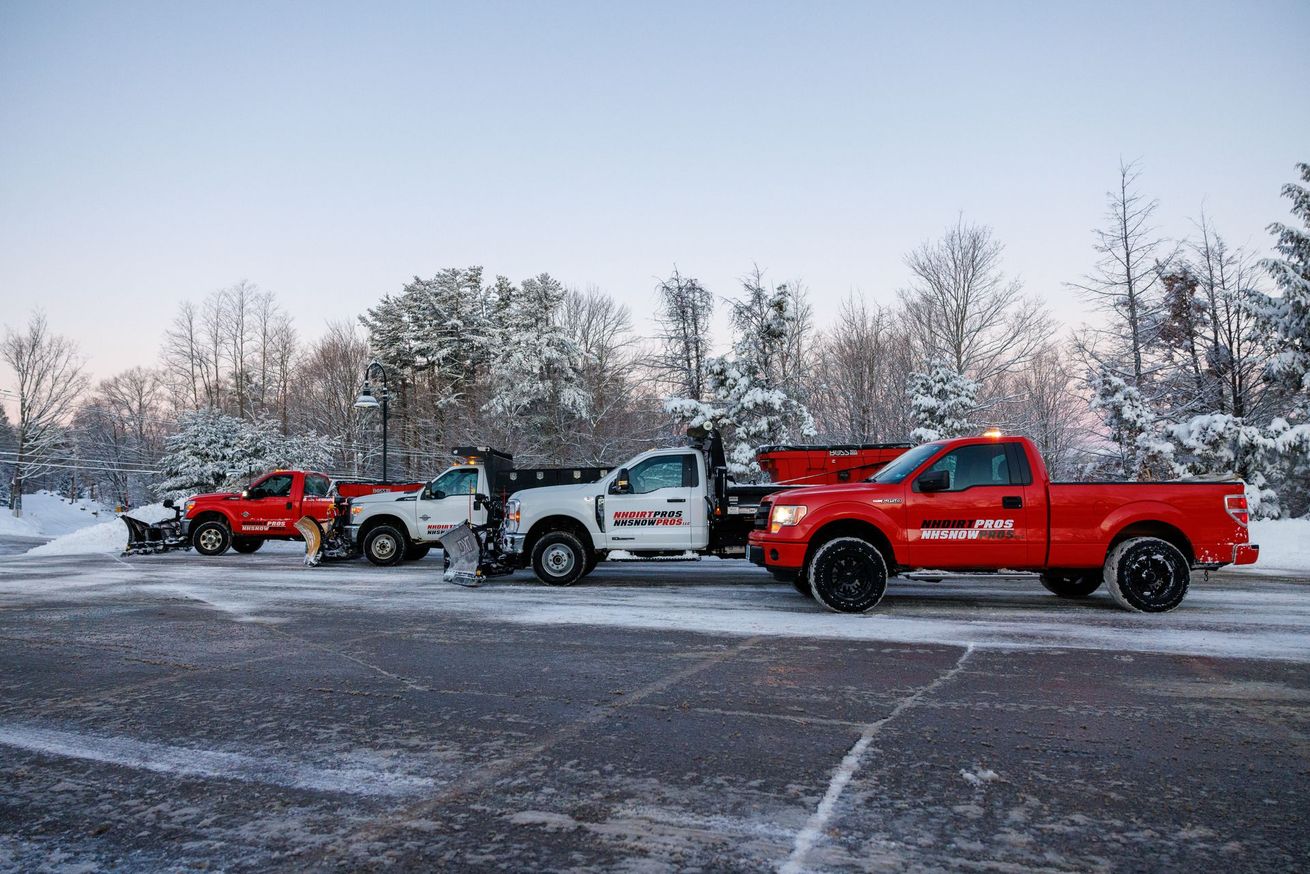 A line of red and white pickup trucks with snow plows parked in a snowy parking lot surrounded by frost-covered trees.