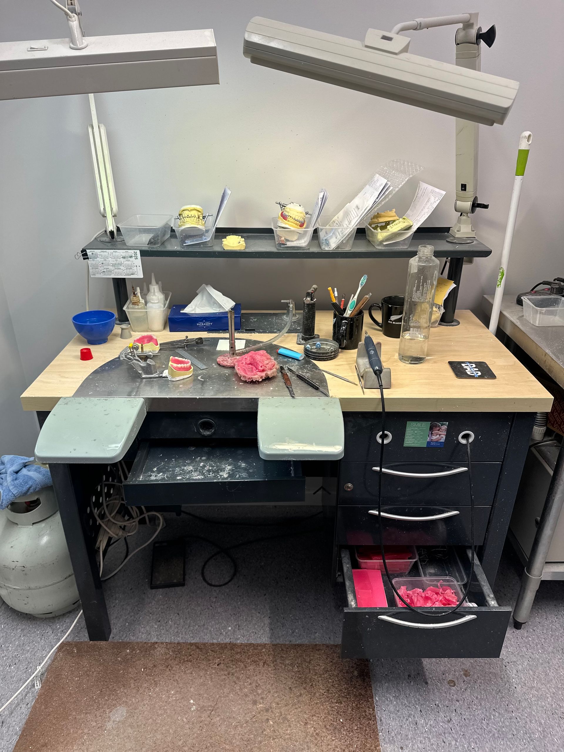 Dental lab workstation with dental models, tools, and overhead lamps. The workstation is gray and has drawers.