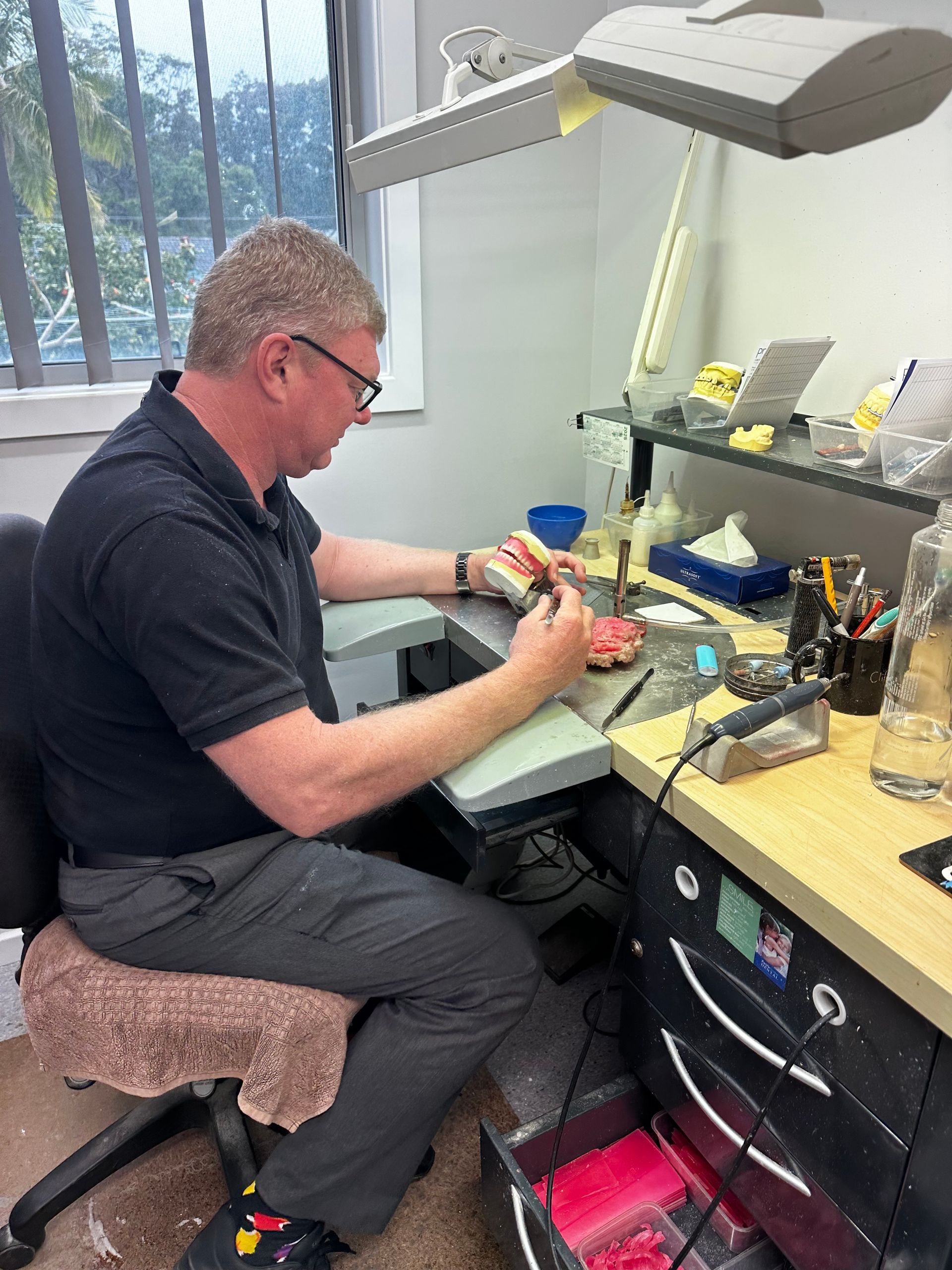 Man in glasses working on dentures at a workbench. He's in a dental lab, using tools with focused concentration.