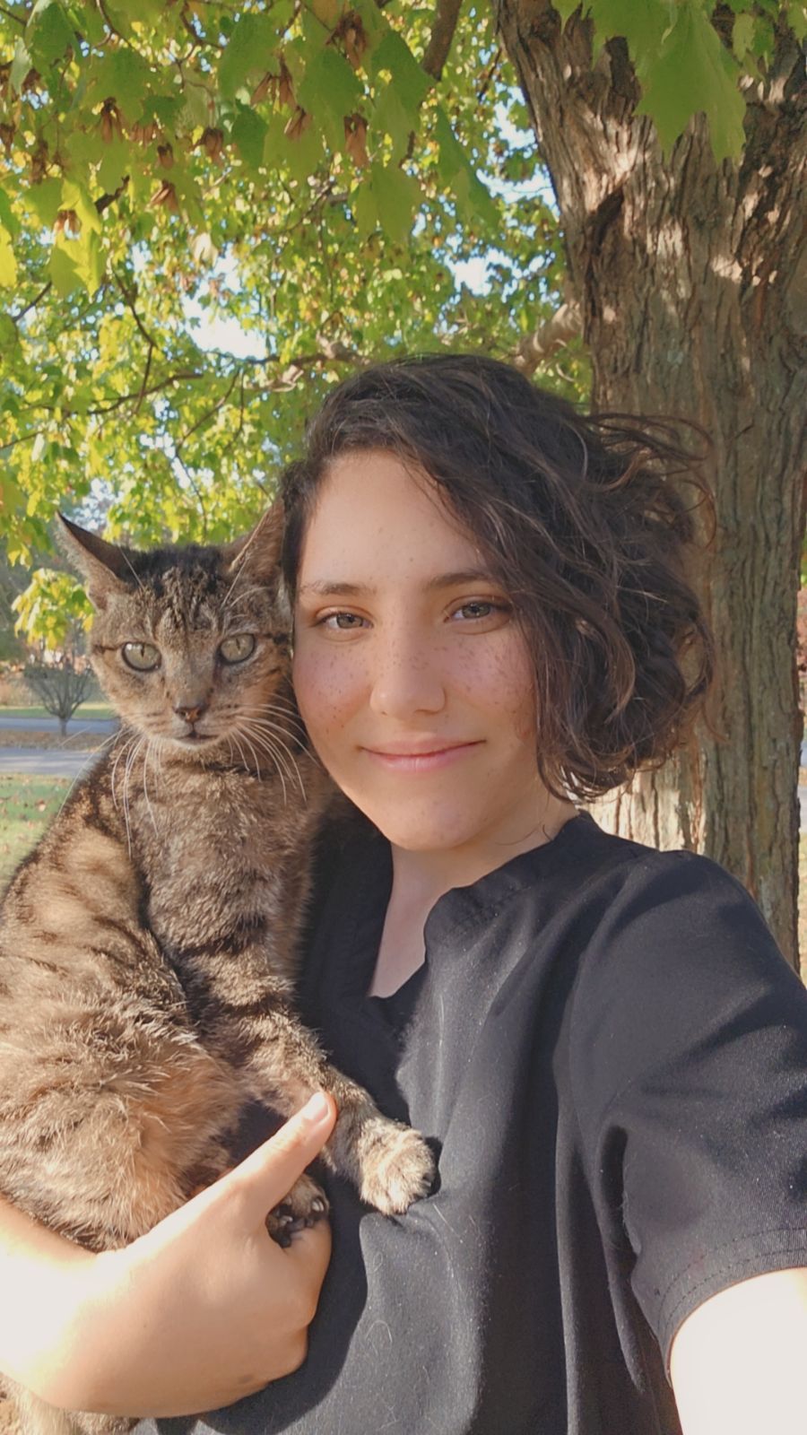A woman is holding a cat in her arms in front of a tree.