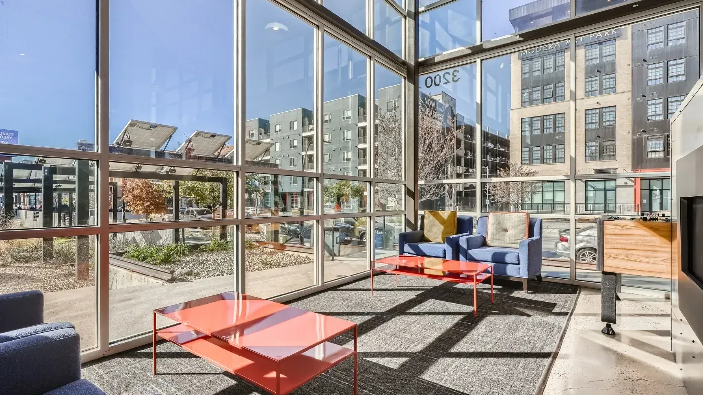Bright communal lounge with floor-to-ceiling windows, blue chairs, and red coffee tables.