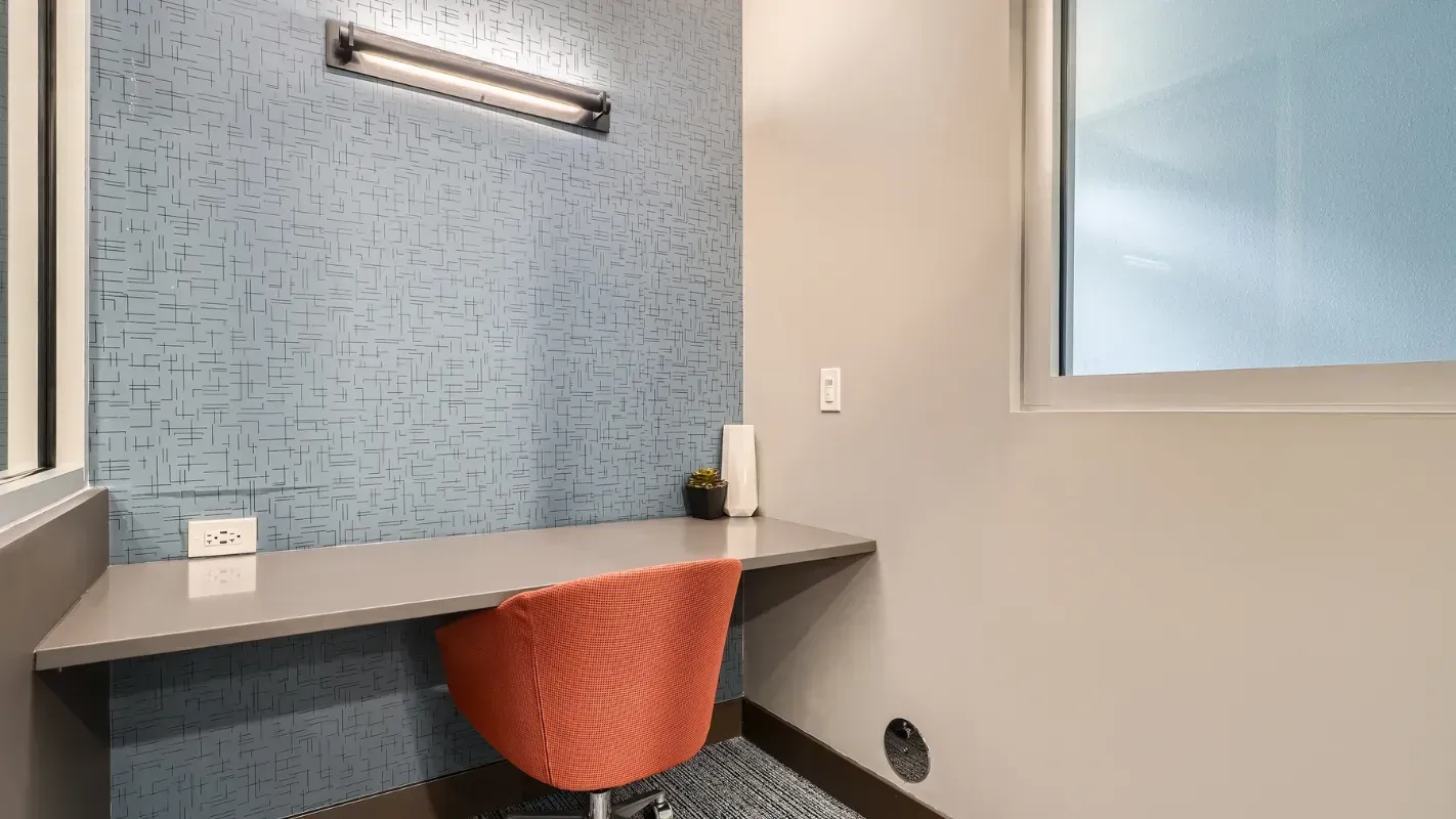 In-apartment workspace with a long grey desk, blue geometric wallpaper, and an orange chair.