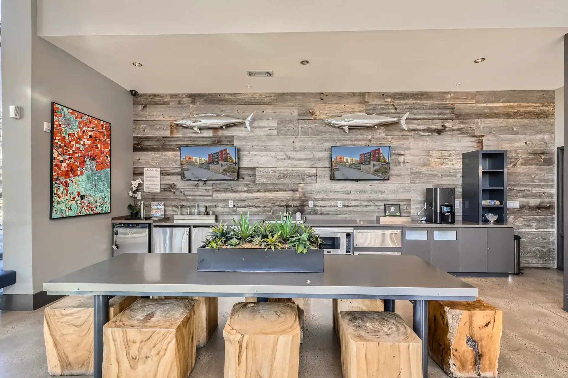 Interior community lounge with a long table, wooden block stools, plants, and a kitchenette against a wood-paneled wall.