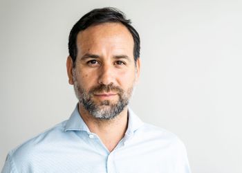 Un uomo con la barba indossa una camicia blu e guarda la telecamera.