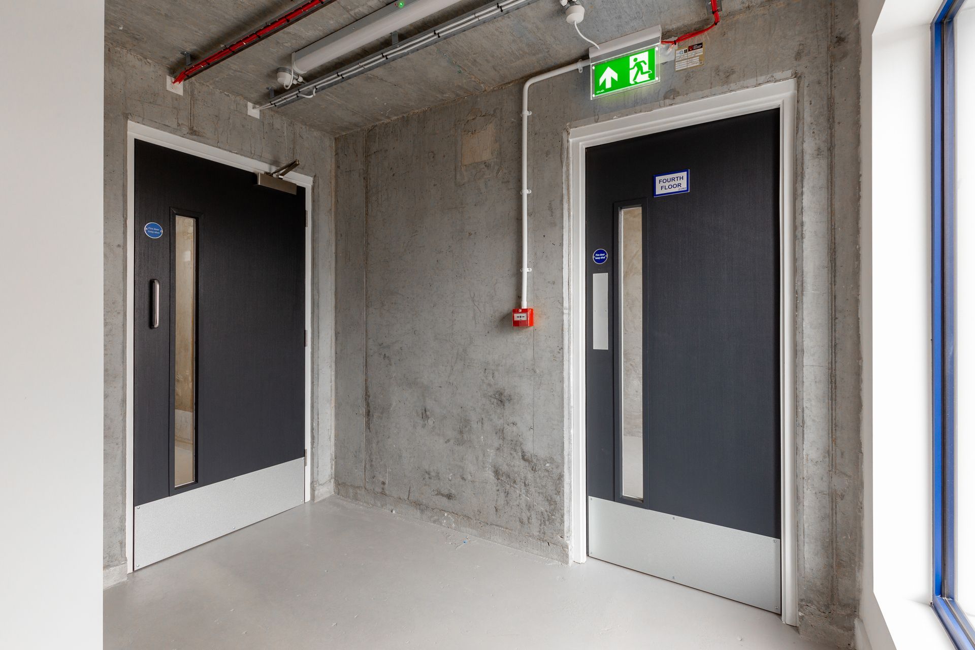 Two fire doors in a concrete hallway with an exit sign and a fire alarm button on the wall.