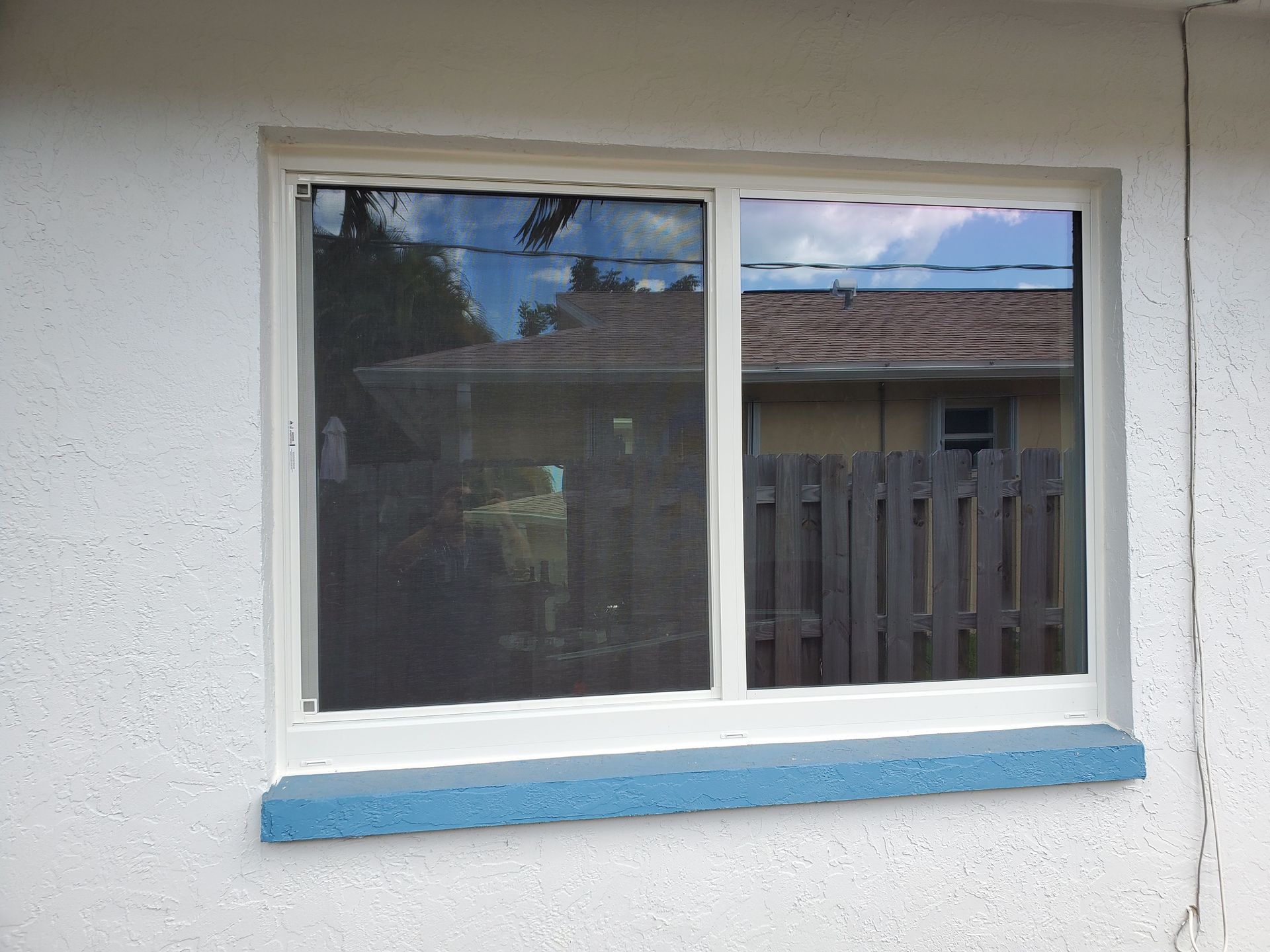 White framed sliding window in a white stucco wall, with blue window sill. Reflections of a house and fence are visible.