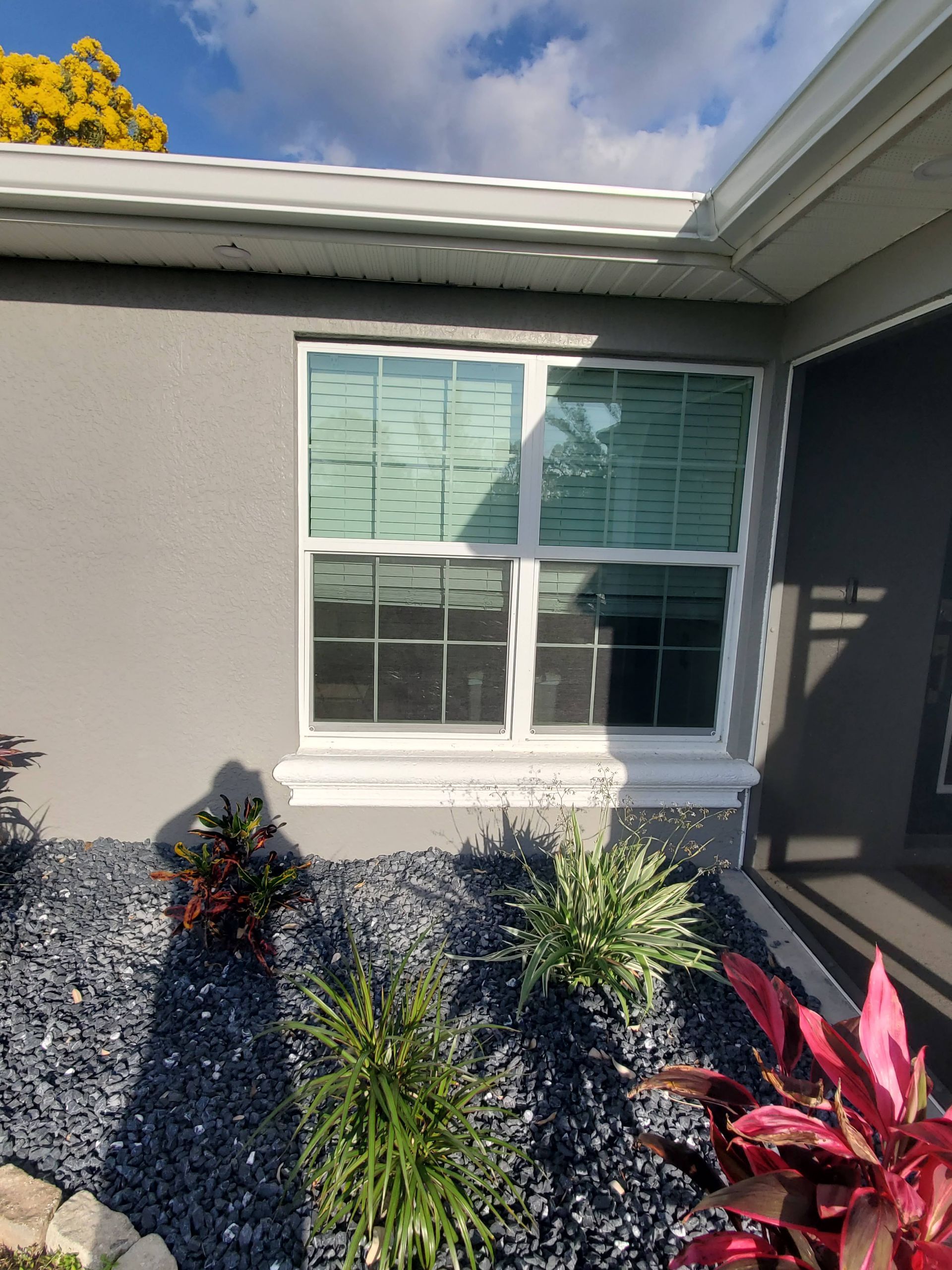 A gray house with a white-framed window; landscaping in the foreground with dark pebbles and plants.