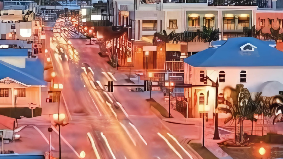 Street scene at dusk with blurred vehicle lights and buildings with blue roofs and palm trees.