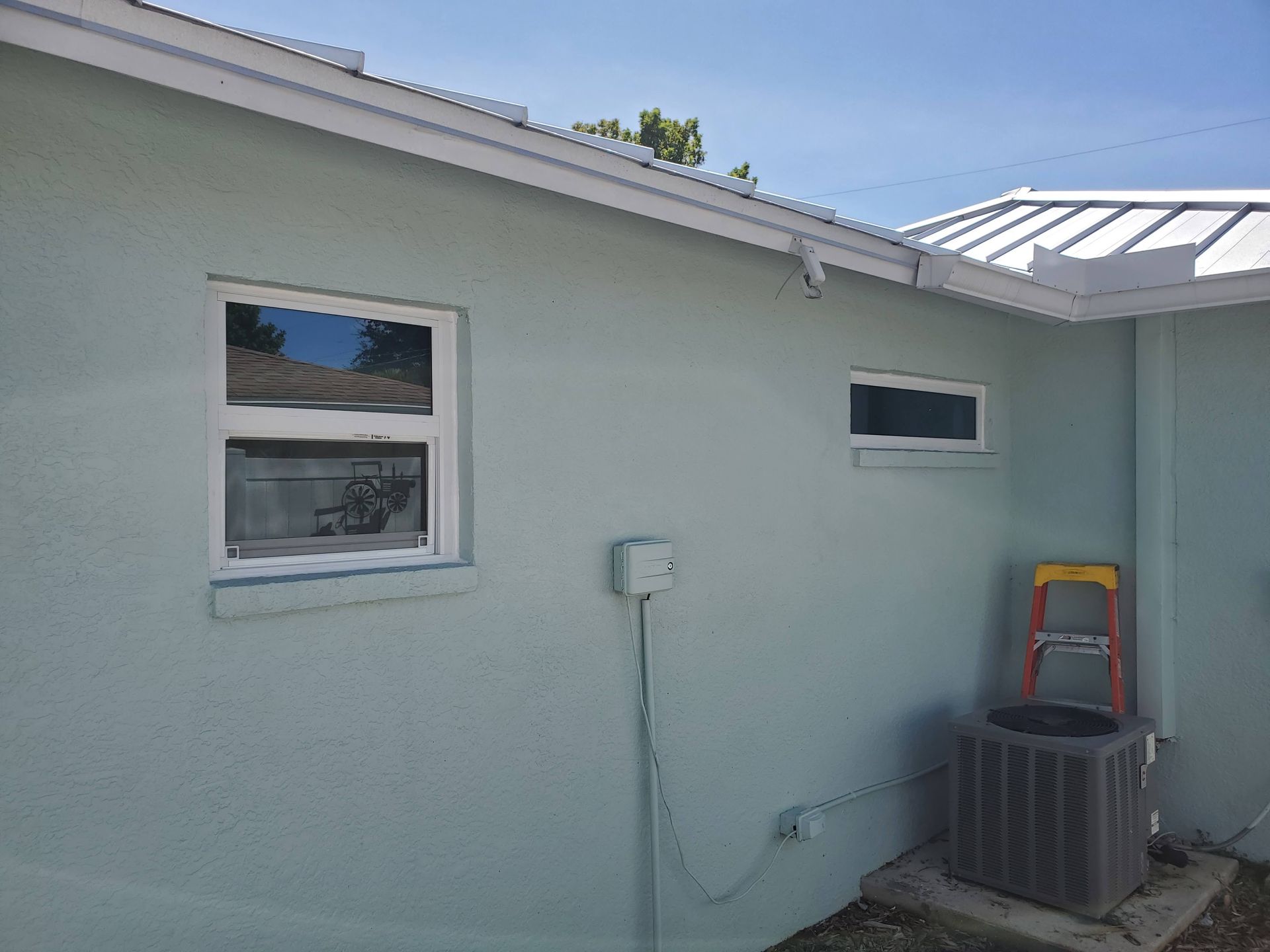 Light blue house exterior with white-framed windows, an air conditioner, and a ladder on a sunny day.