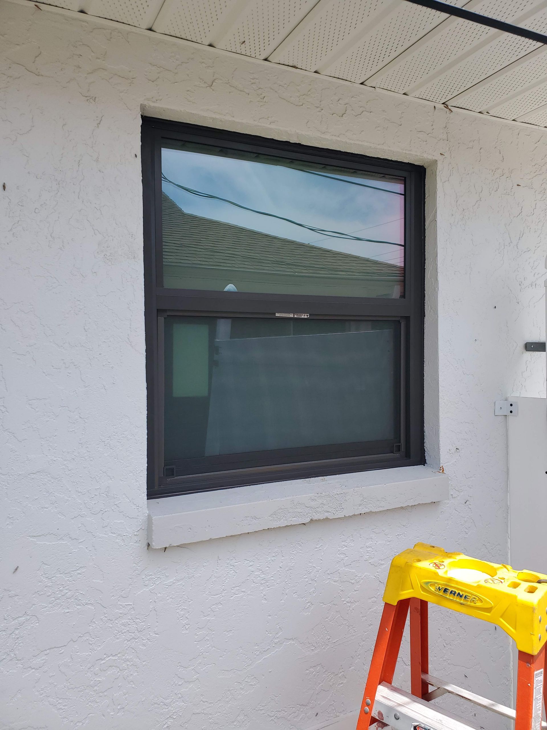 Black framed window on a white stucco wall. A yellow ladder is visible at the right.