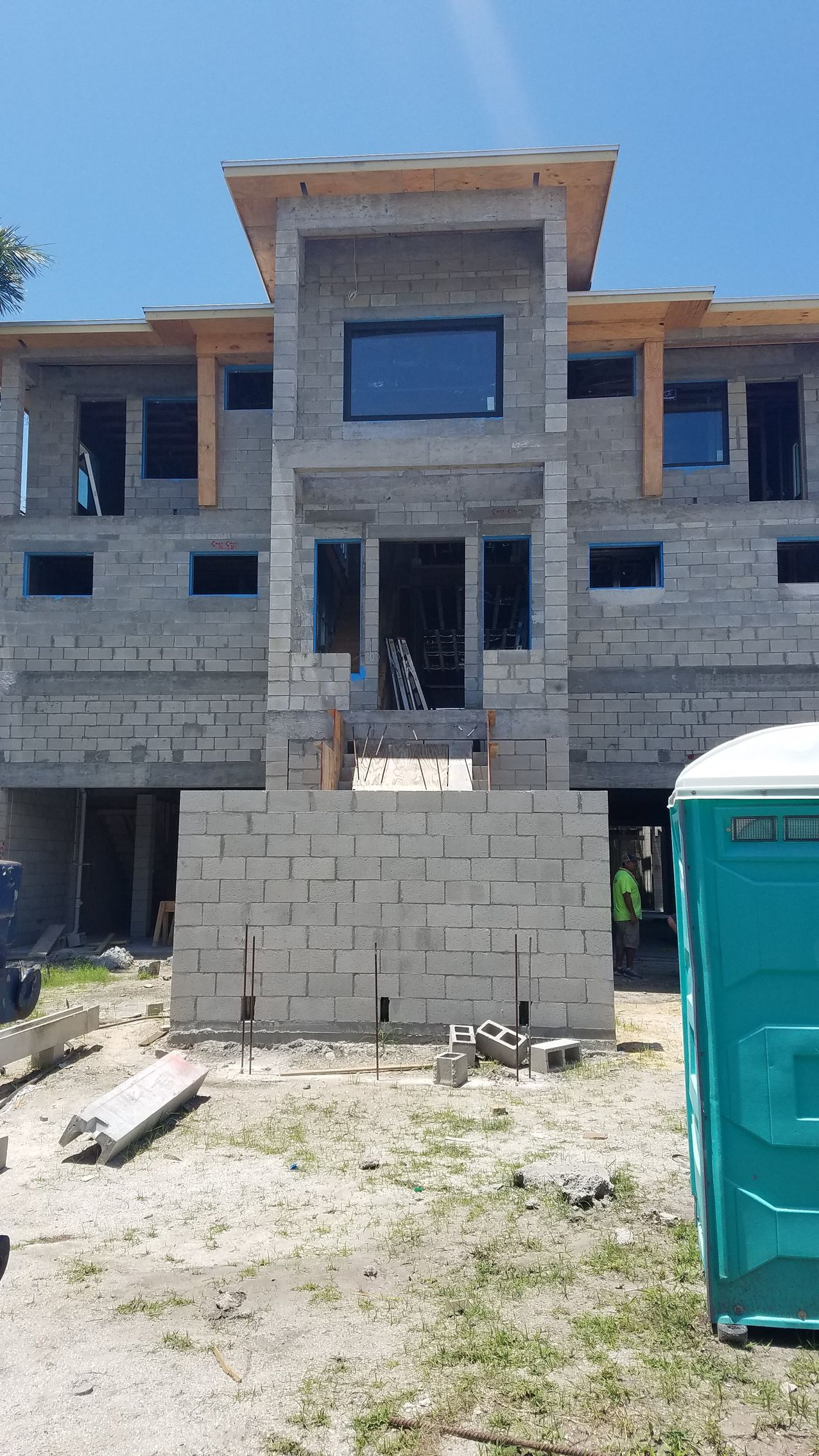 A partially constructed two-story gray block building with windows, surrounded by construction materials, and a porta-potty.