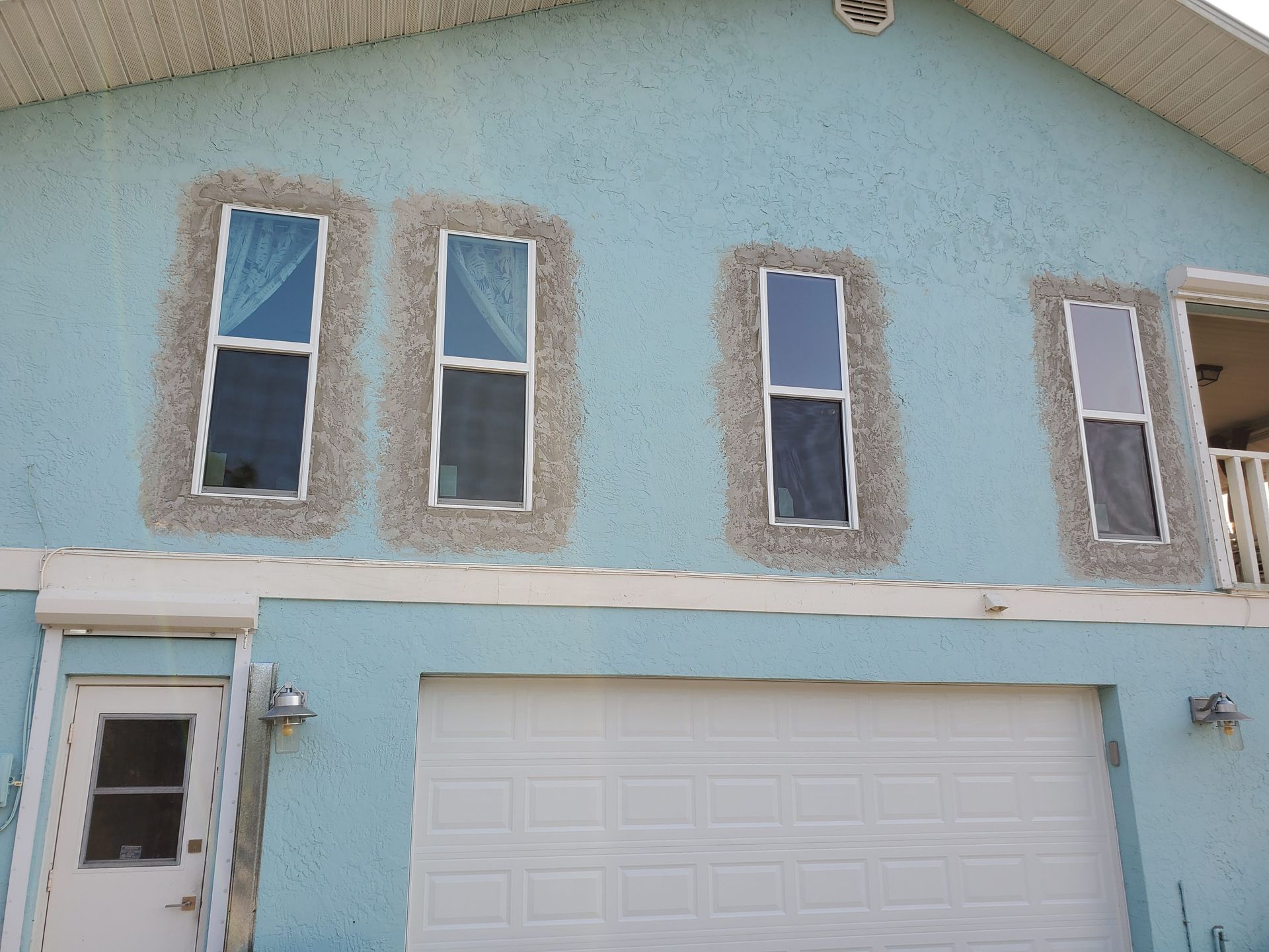 Blue house exterior with four windows framed in rough stucco; a garage door and front door are visible.