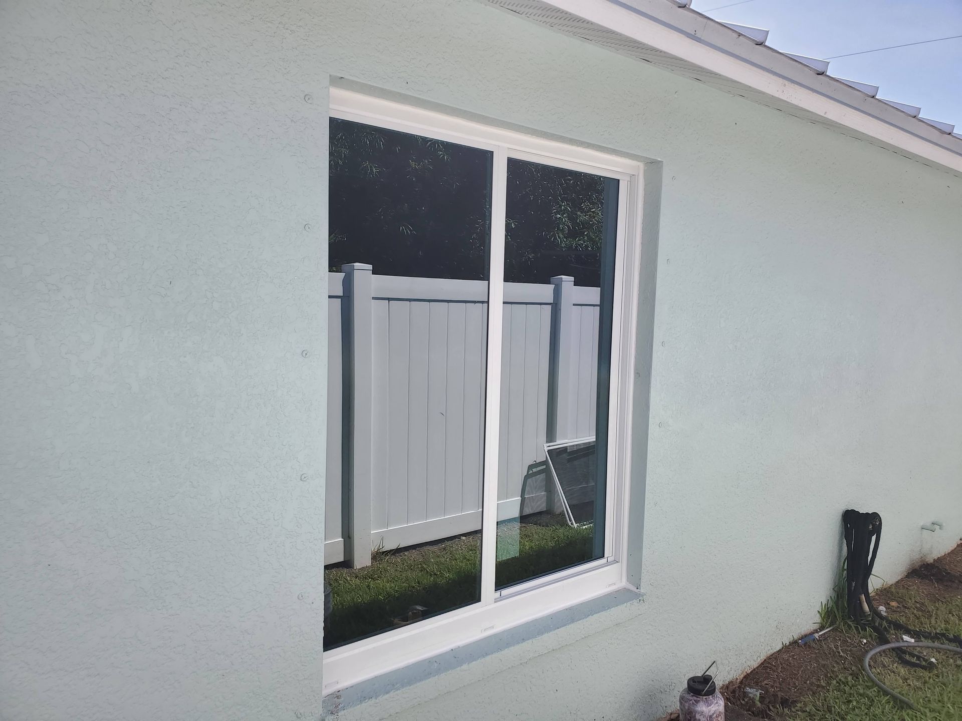 White-framed sliding glass window on a light blue house, reflecting a white fence and grass.