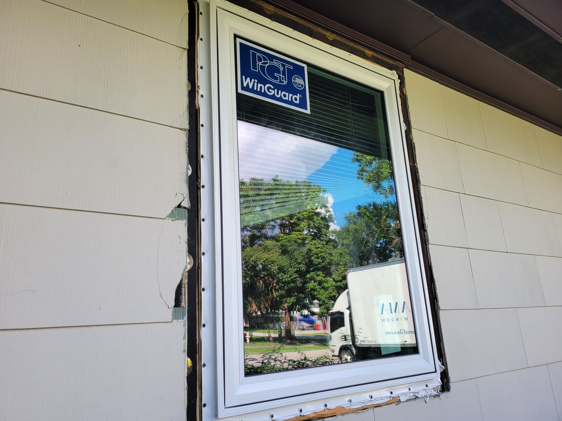 White framed window installed on a beige sided house, reflecting trees and a white truck.
