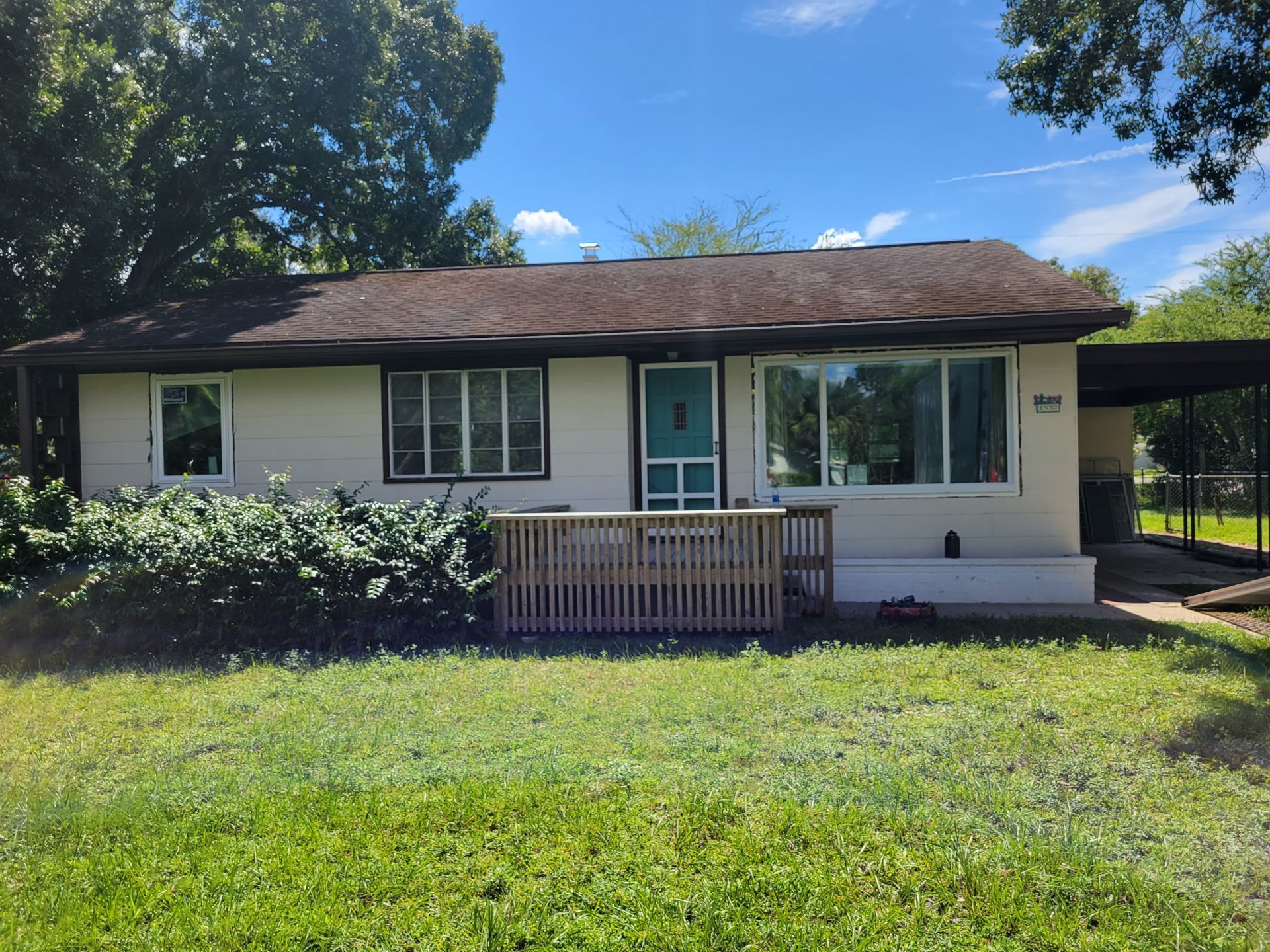 Tan house with turquoise door, wooden porch, and carport. Green grass and trees in the sunny yard.