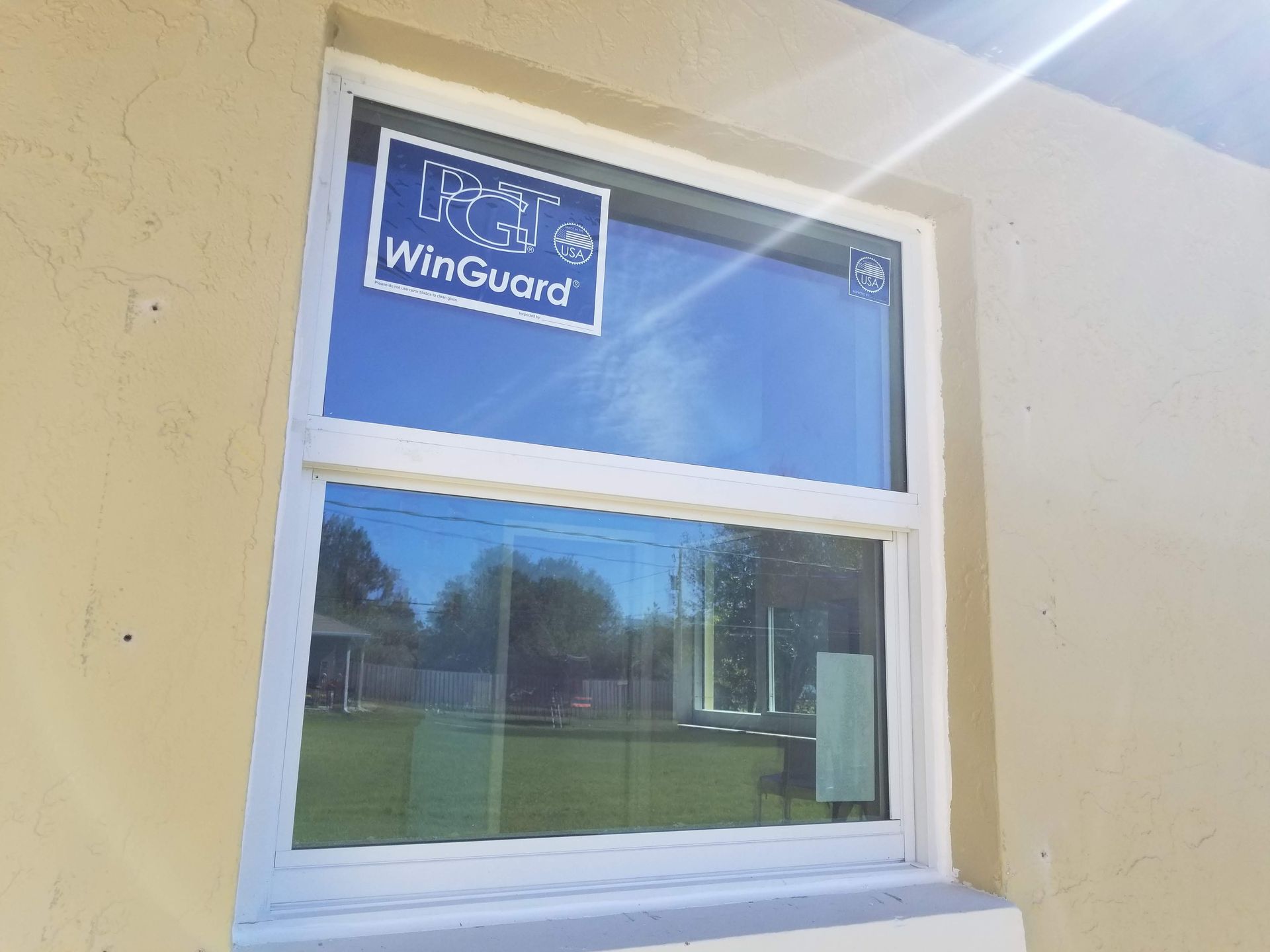 White-framed double-hung window in a light beige wall, reflecting a blue sky and green lawn.