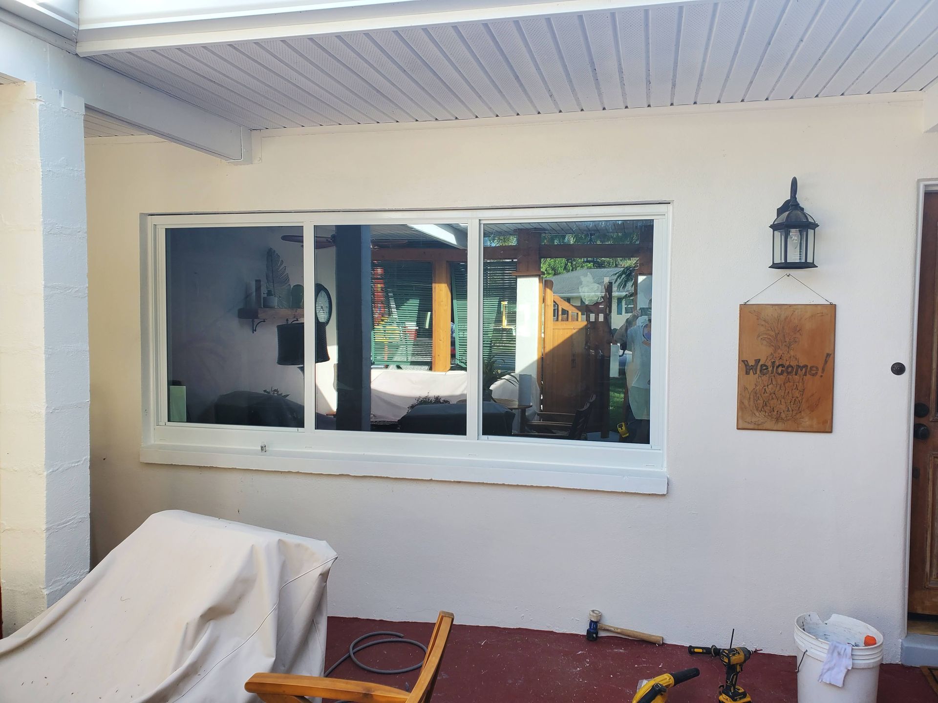 Exterior view of a house with a large window and an overhead porch. Sunlight and a brown wooden door are visible.